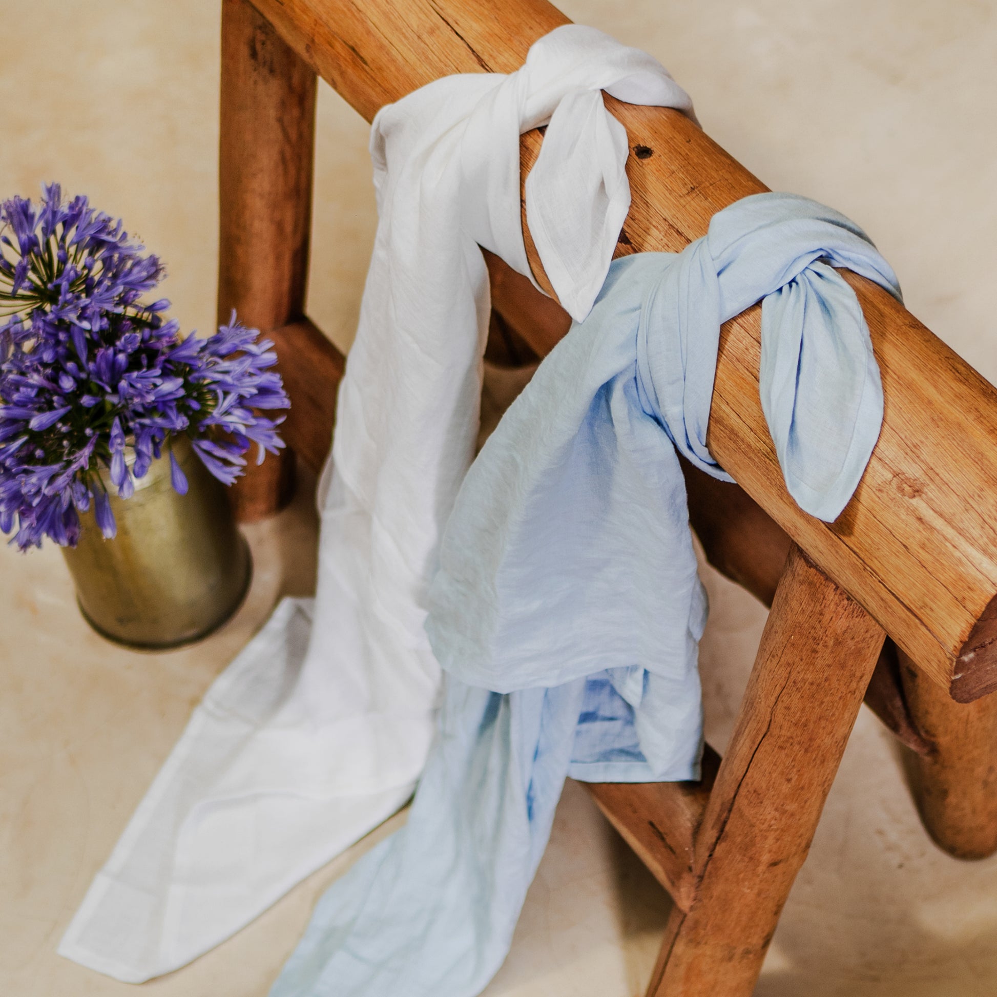 Two linen swaddles, one white and one blue, are draped over a wooden bench with a vase of purple flowers beside them.