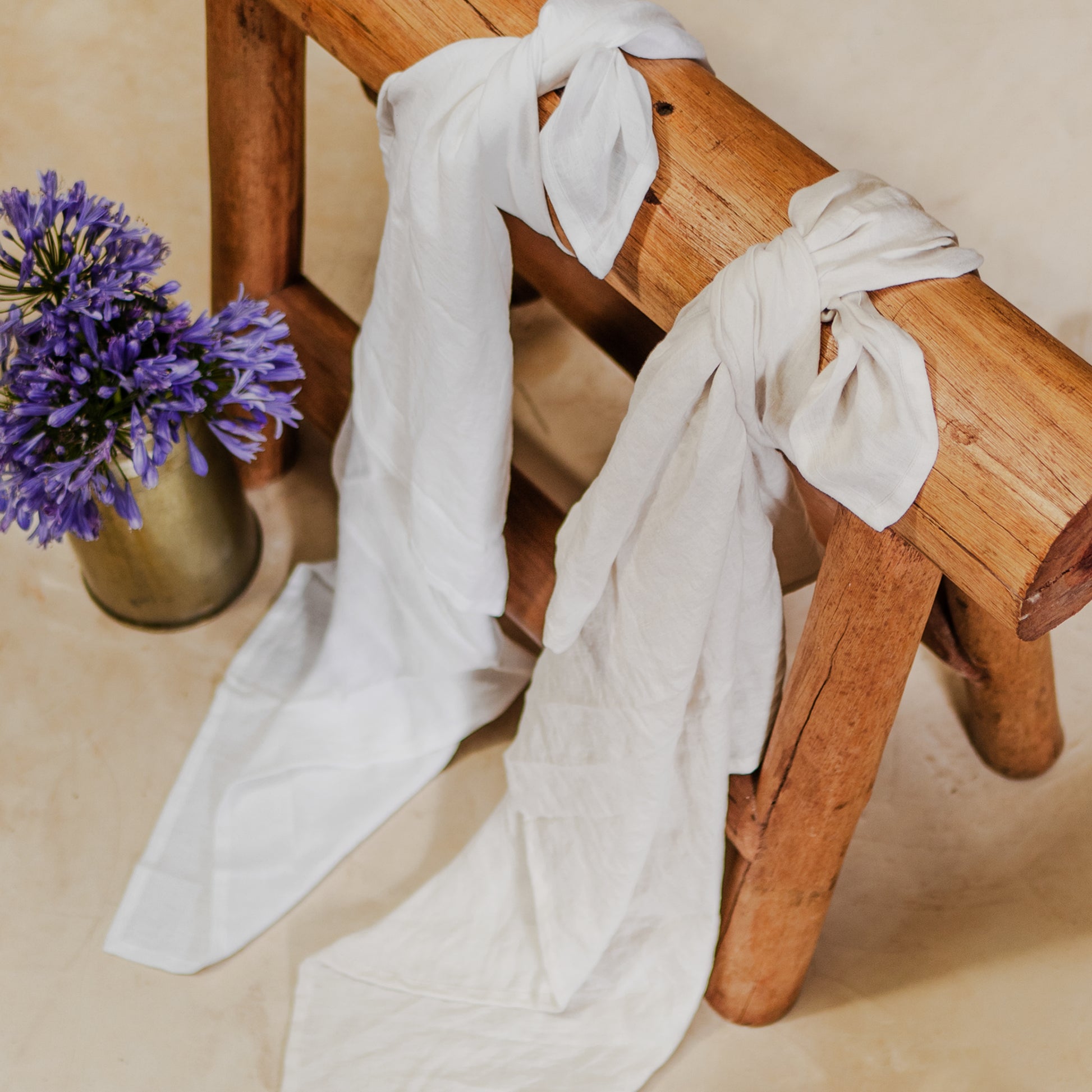 Two linen swaddles, one white and one blue, are draped over a wooden bench with a vase of purple flowers beside them.